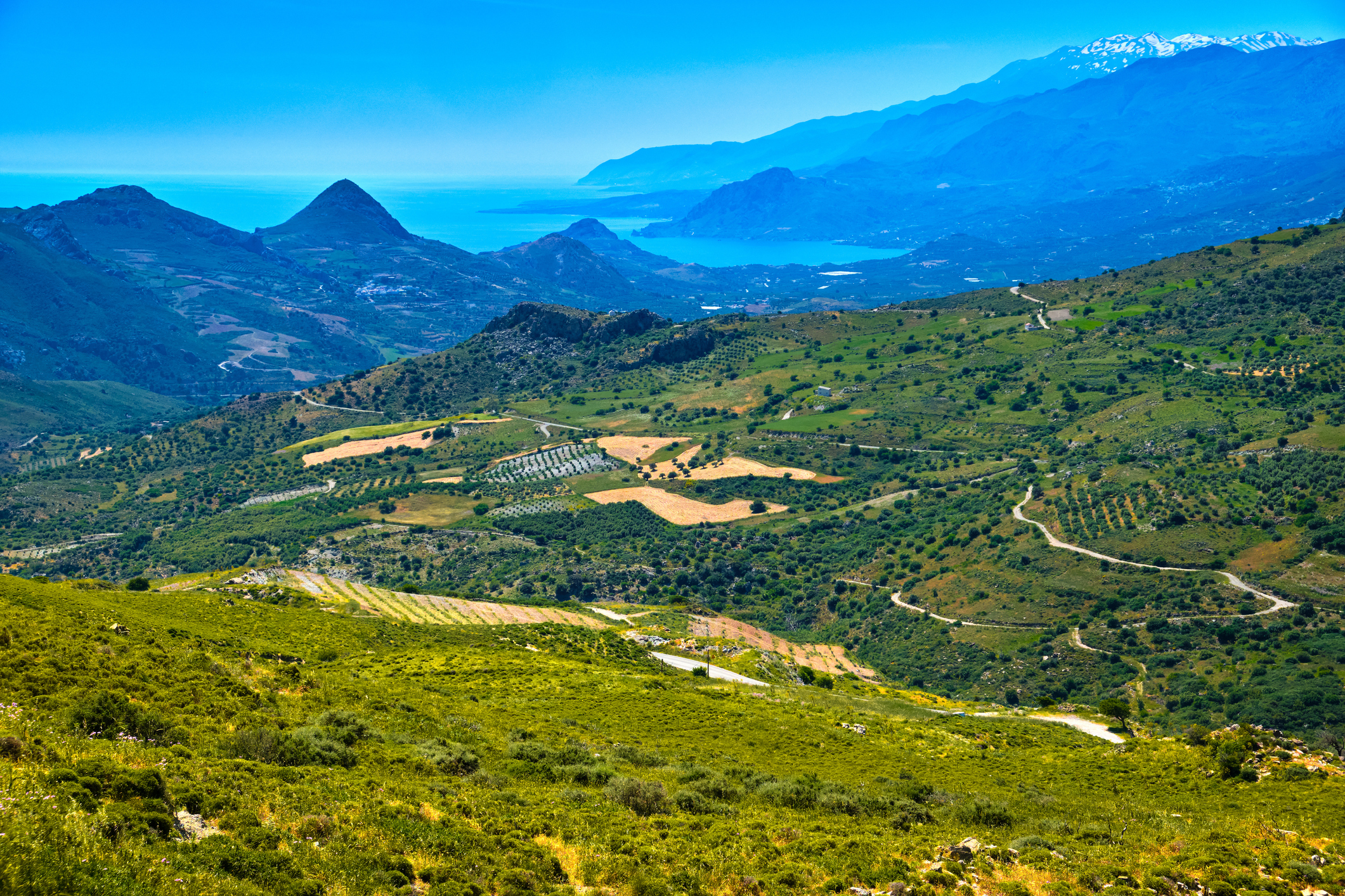 Summer day landscape of Crete island, Greece. High mountains, snow-covered peaks, green hills, roads and olive tree plantations, high grass, lush foliage and vegetation. Mediterranean sea, blue sky and clouds. Idyllic panorama on sunny day.